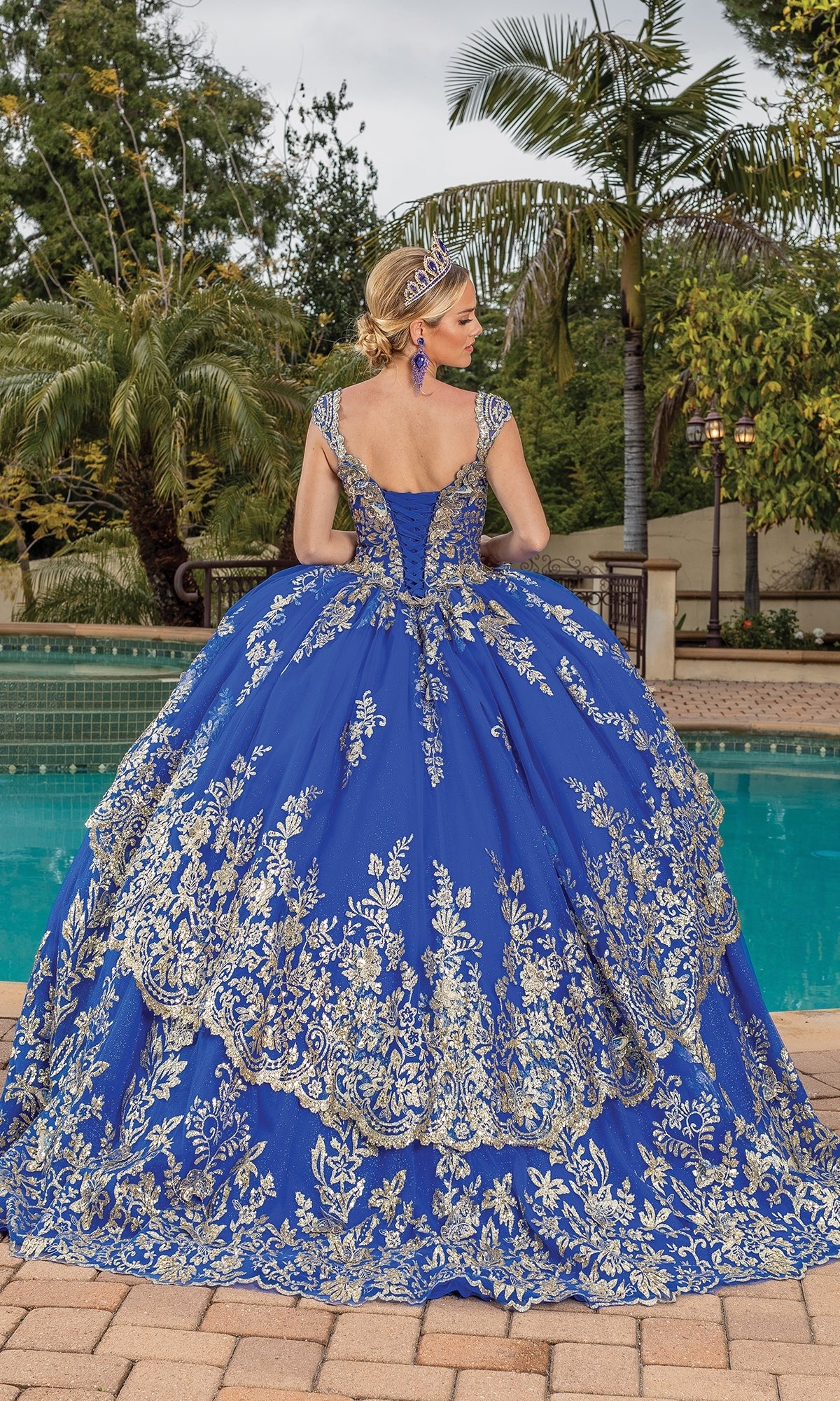 A woman wearing the Quinceanera Dress 1881 By Dancing Queen in royal blue with gold lace applique stands by a pool, facing away, surrounded by palm trees and lush greenery.